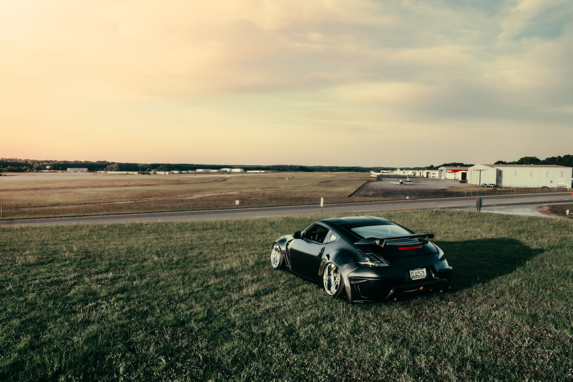 Black widebody Nissan 370Z parked on a grass hill beside an airport at sunset