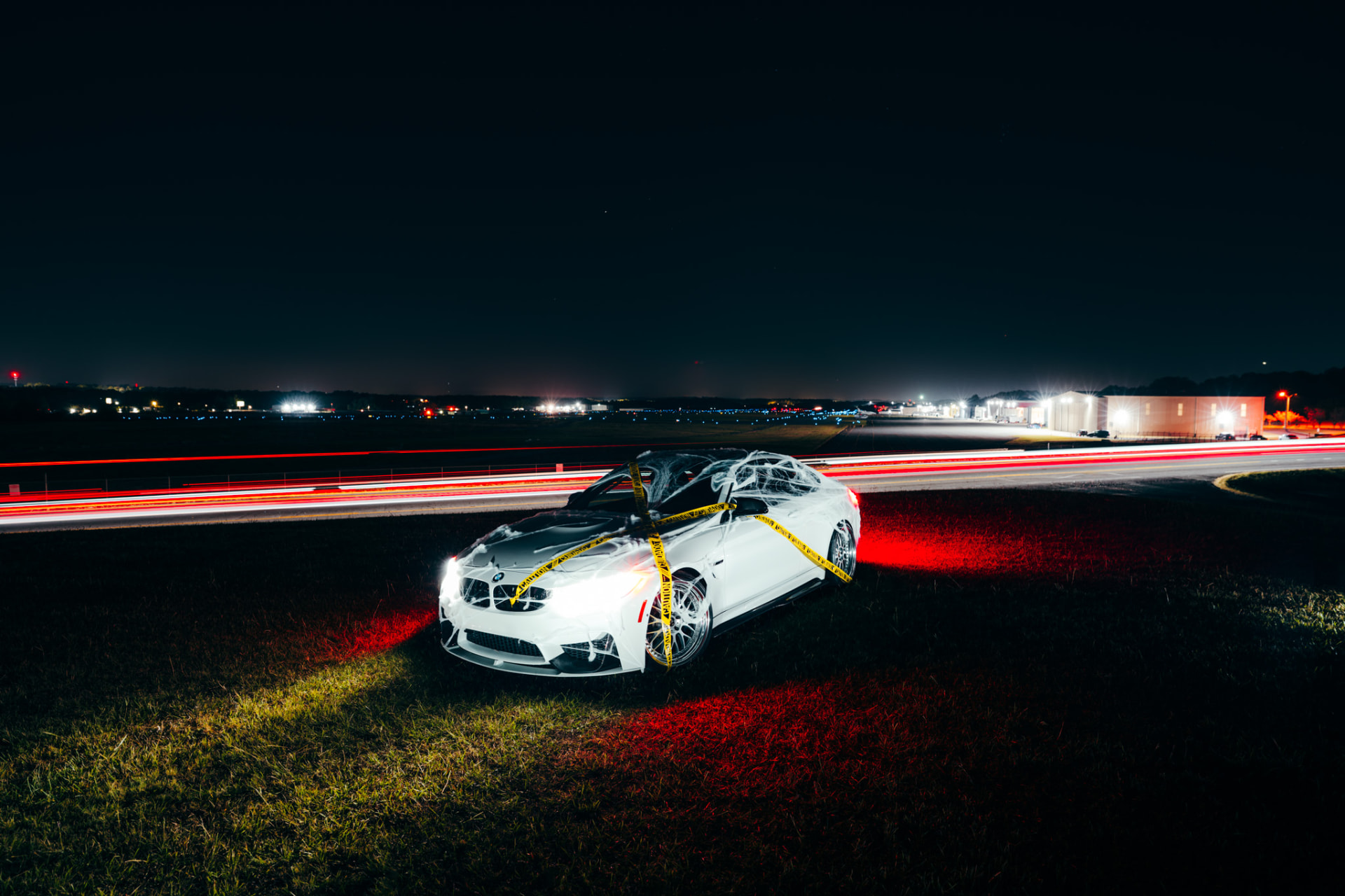 White BMW M4 wrapped in cobwebs and caution tape beside airport runway lights at night